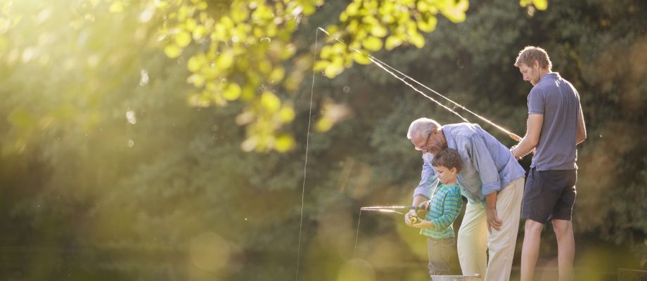 Son, pappa och farfar metar tillsammans vid en å - bilden är tagen på avständ med grönska som delvis döljer bilden. En stillsam eftermiddag med familjen.